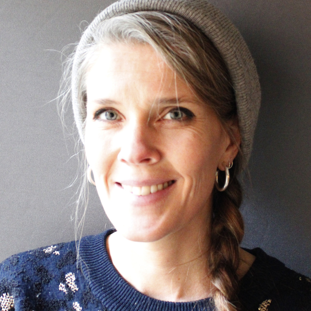 Leende kvinna med fläta, grå mössa och örhängen mot en enfärgad bakgrund.