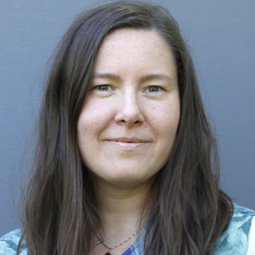 Kvinna med långt brunt hår som ler lätt, klädd i en blå topp, framför en grå bakgrund.