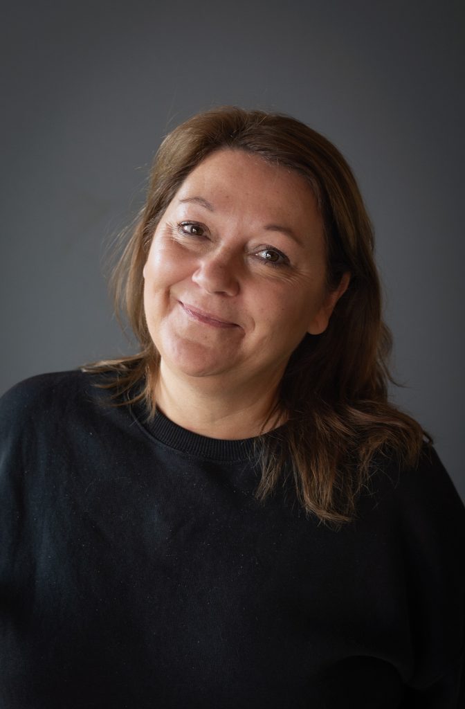 En kvinna med brunt hår och svart topp ler varmt mot en vanlig mörk bakgrund.