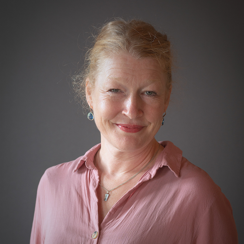 Leende kvinna med blont hår i rosa blus, stående mot en grå bakgrund.