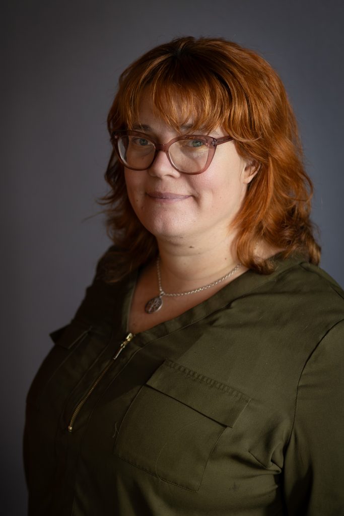Rödhårig kvinna med glasögon, olivgrön topp och ett litet leende mot en grå bakgrund.