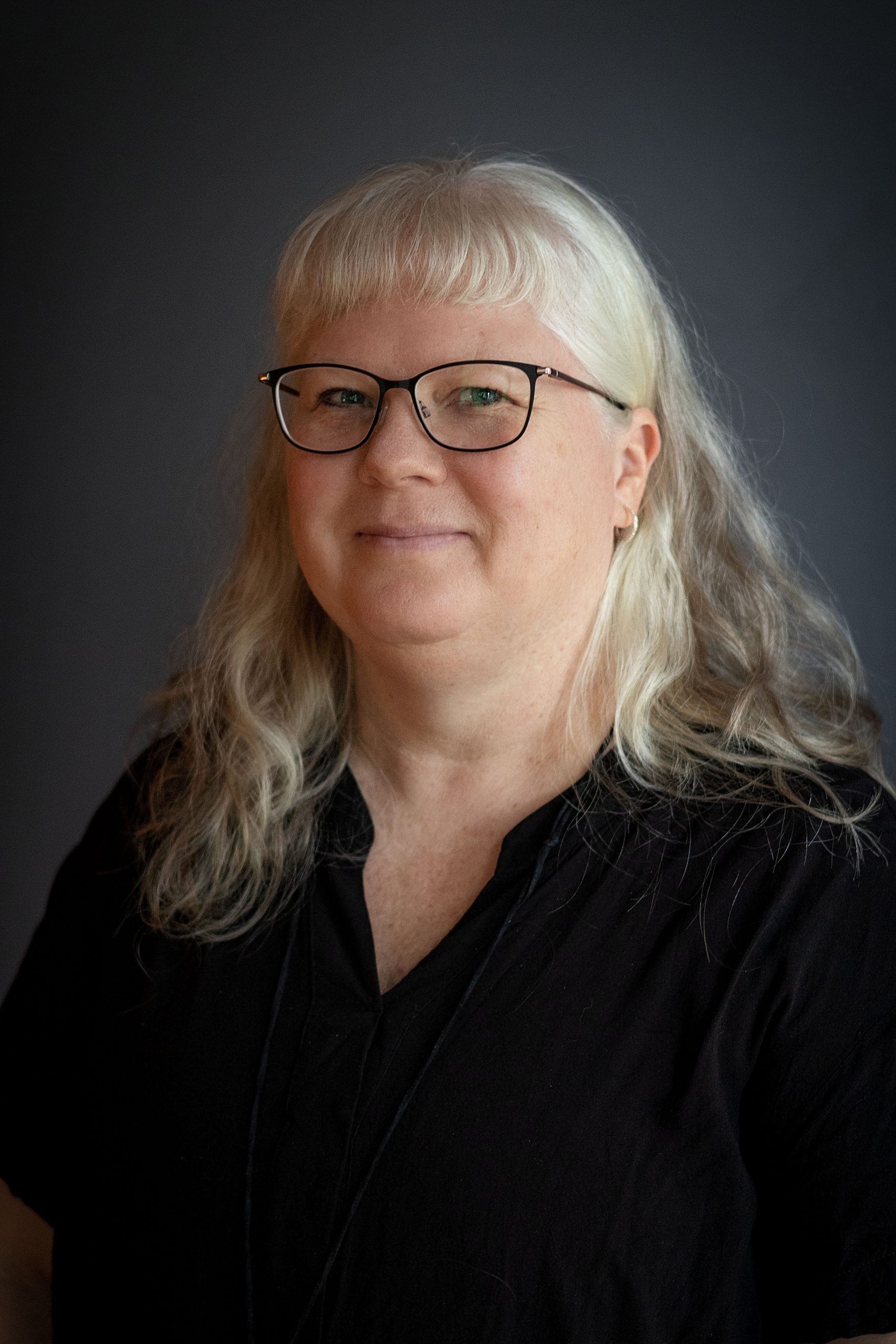 Leende kvinna med långt grått hår, iförd glasögon och svart skjorta, mot mörk bakgrund.
