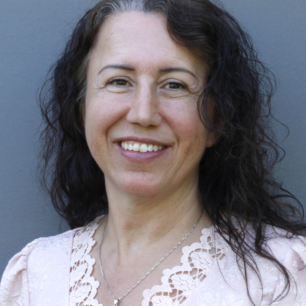 Leende kvinna med vågigt mörkt hår, klädd i en ljusrosa spetstopp, mot en grå bakgrund.