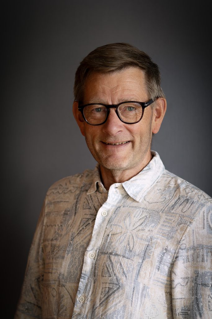 Leende äldre man med glasögon och mönstrad skjorta poserar mot en enfärgad, mörk bakgrund.