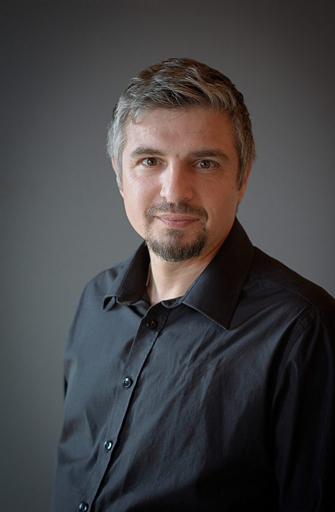 En man med kort grått hår och getskägg, klädd i svart skjorta, står mot en enfärgad bakgrund.