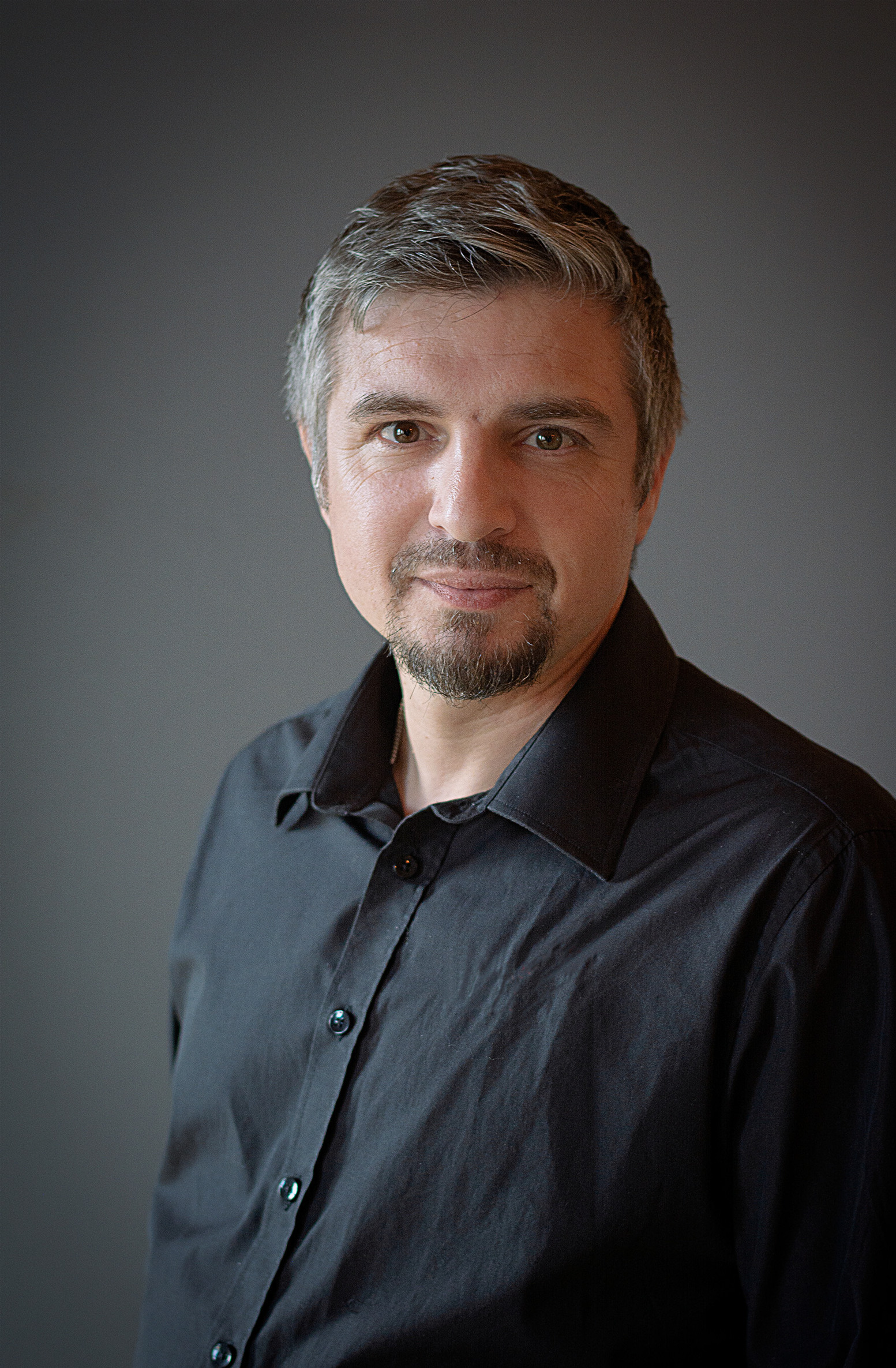 En man med kort grått hår och getskägg, klädd i svart skjorta, står mot en enfärgad bakgrund.