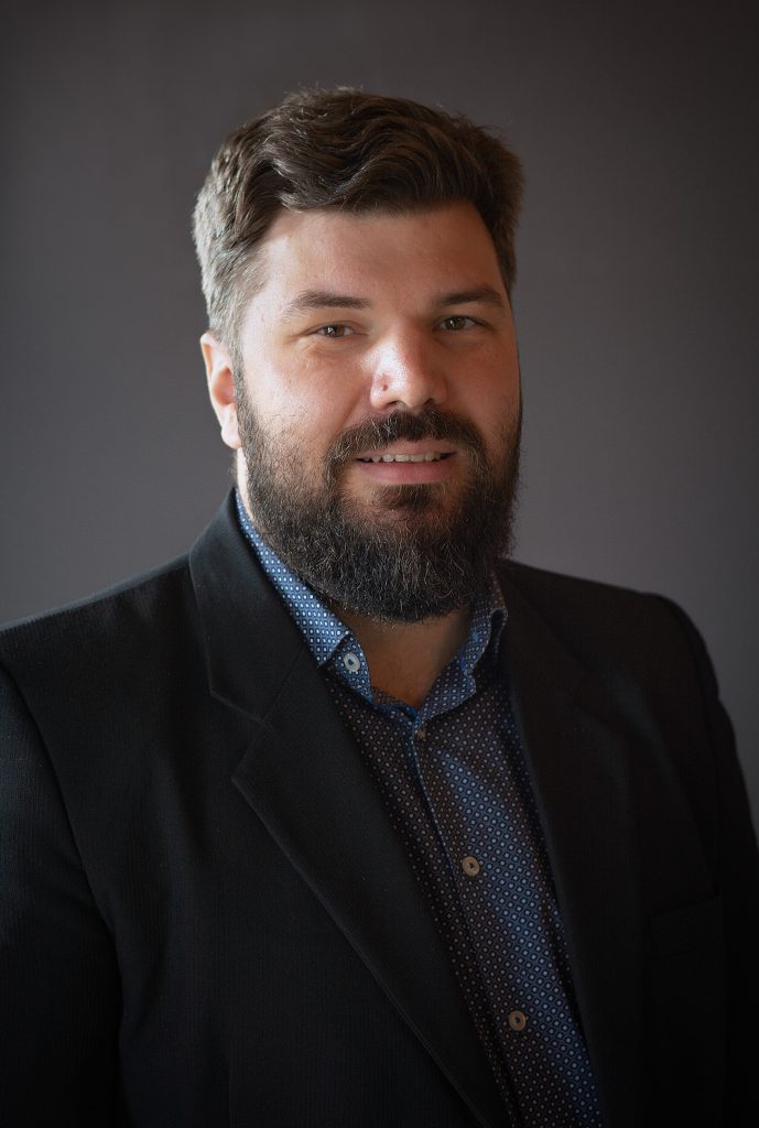 En man med mörkt hår och skägg bär en svart kavaj och blå skjorta och ler mot kameran.