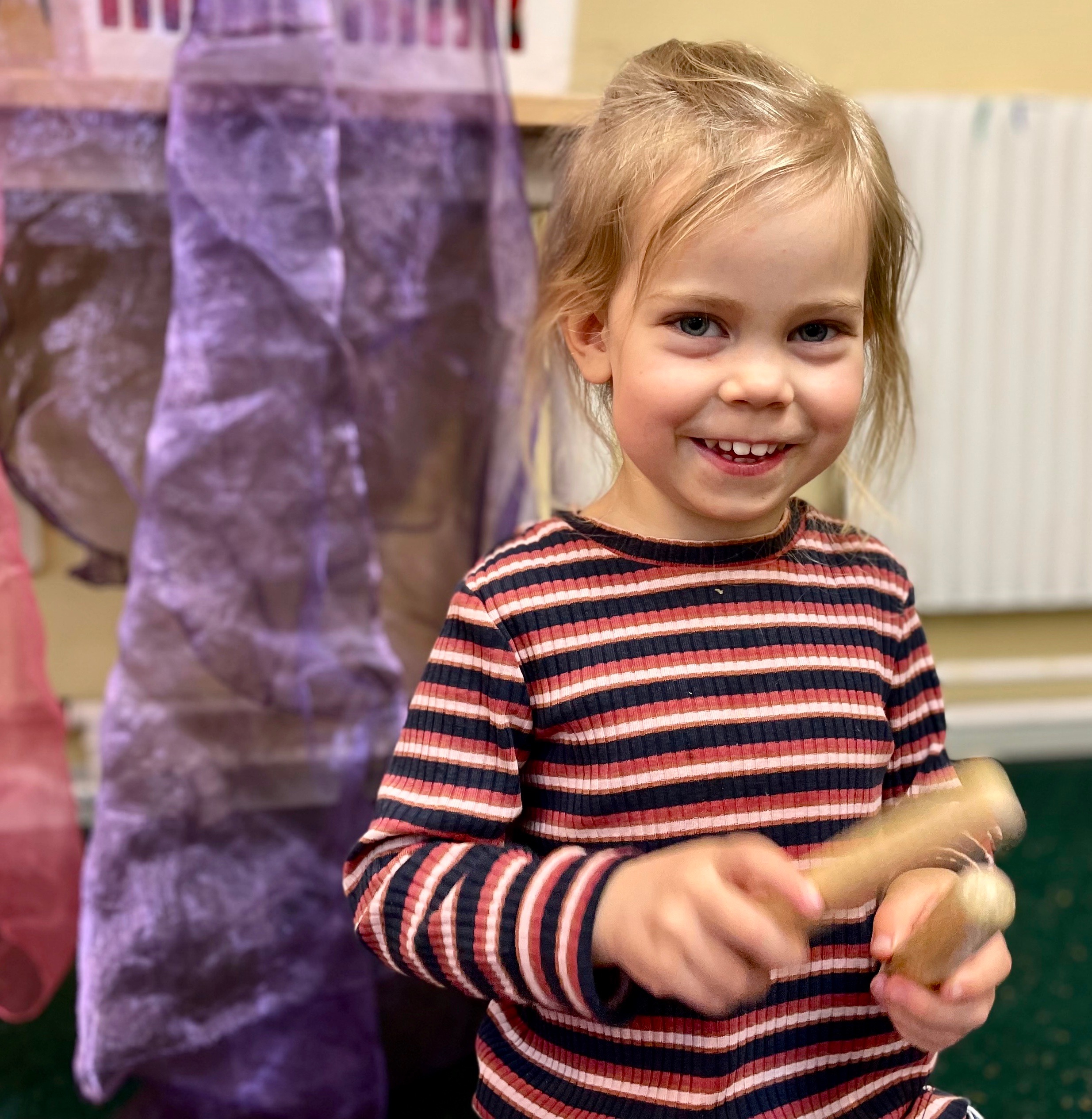 Leende ung flicka i randig skjorta leker med rytmstavar av trä inomhus.