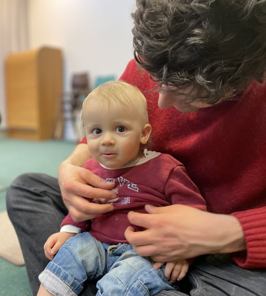 En vuxen i röd tröja sitter med benen i kors och har en bebis i rödbrun skjorta och jeans i knät.