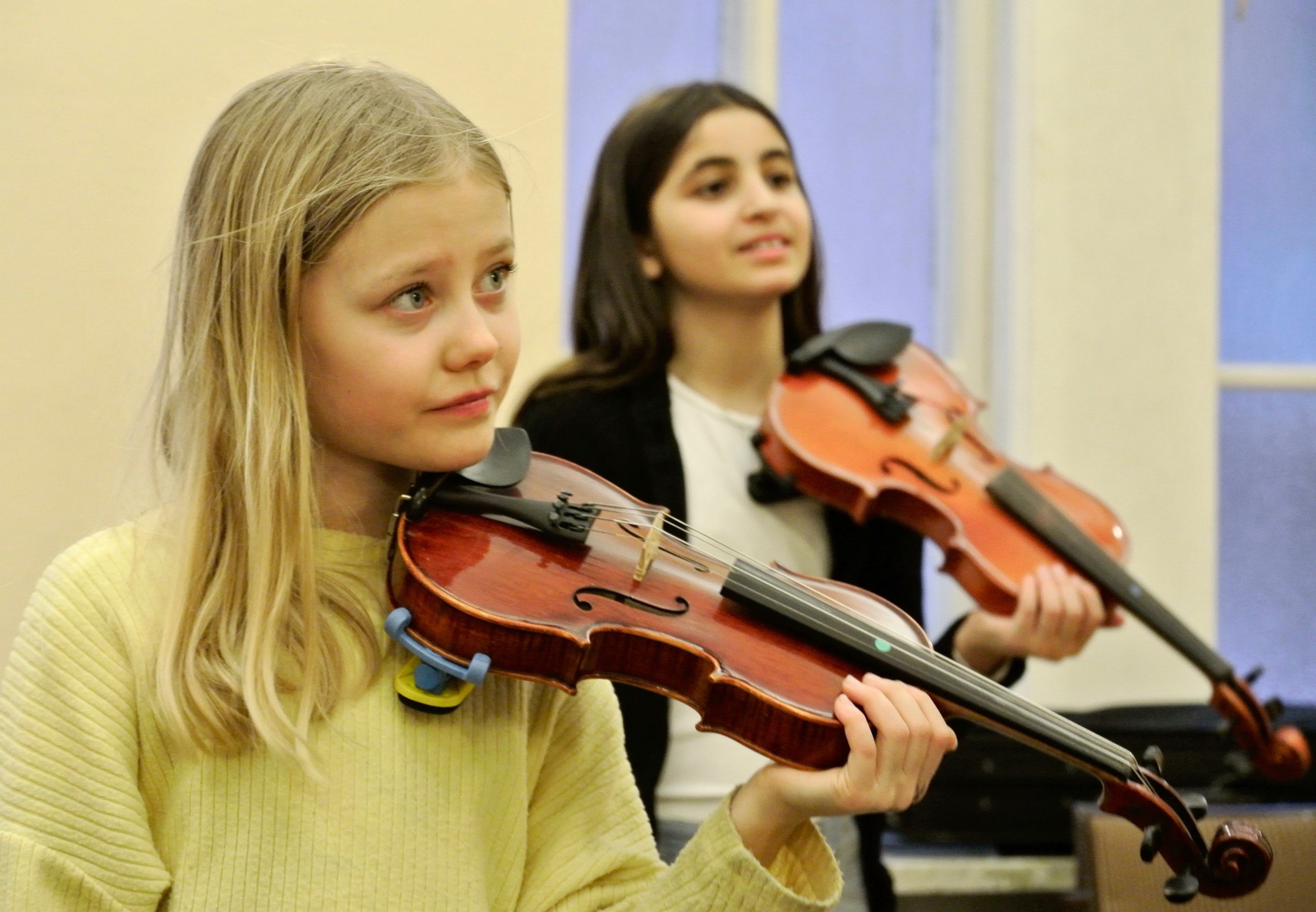 Två flickor spelar violin inomhus och fokuserar på sina instrument med mjukt, naturligt ljus i bakgrunden.