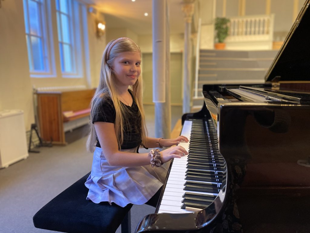 En ung flicka sitter vid en flygel, leende, med händerna på tangenterna i ett starkt upplyst rum.