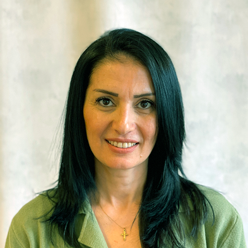 En person med långt mörkt hår, klädd i en grön topp och ett halsband med ett korshänge, ler mot kameran mot en ljus bakgrund.