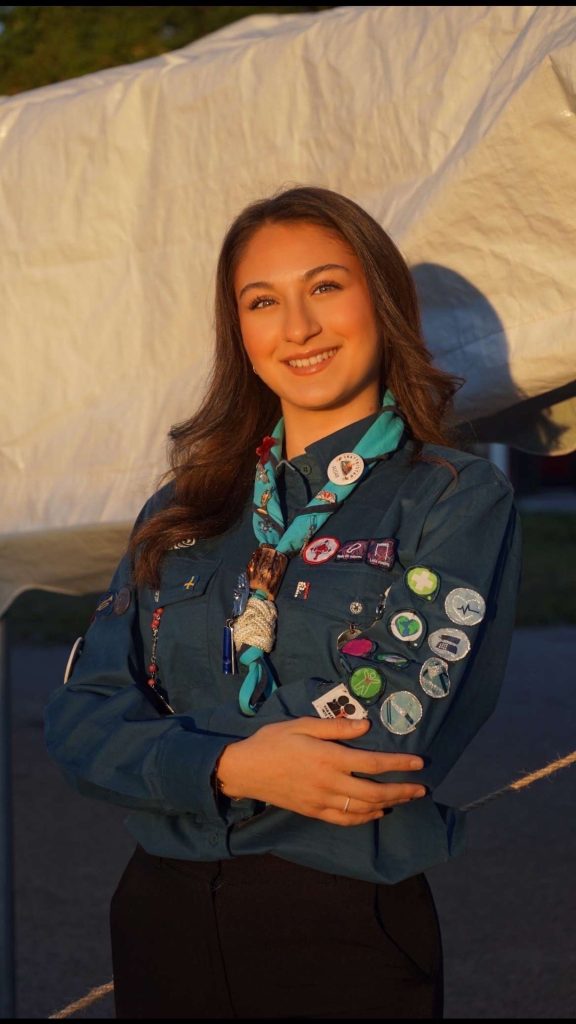 Ung kvinna i scoutuniform med flera märken och pins står utomhus och ler självsäkert med armarna i kors.