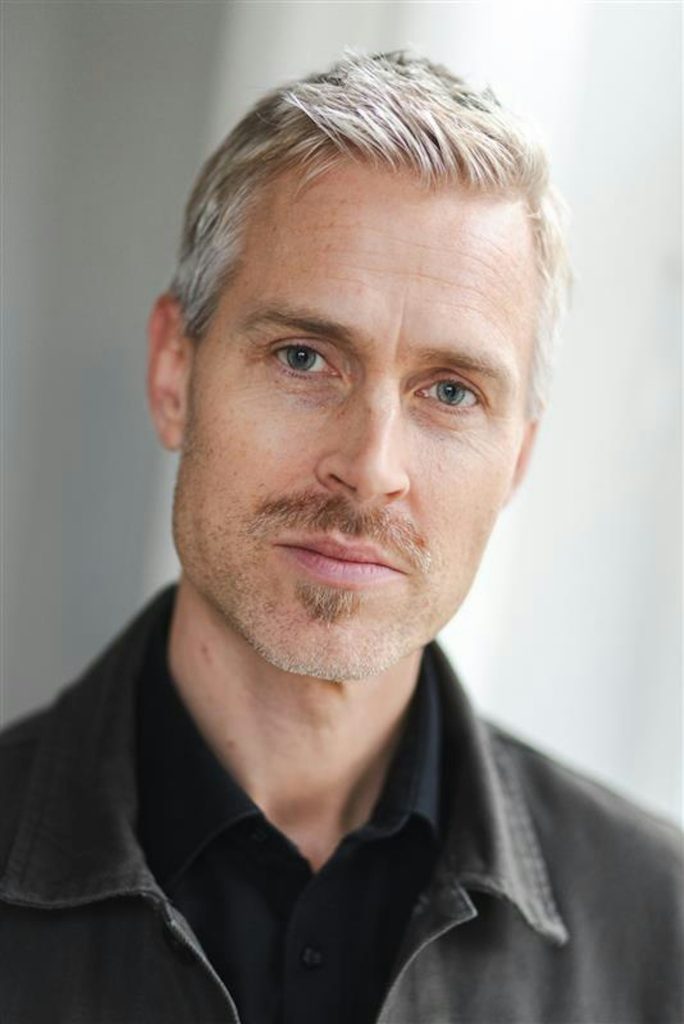 Man med kort grått hår, ljust skägg och blå ögon, klädd i mörk jacka, tittar in i kameran.