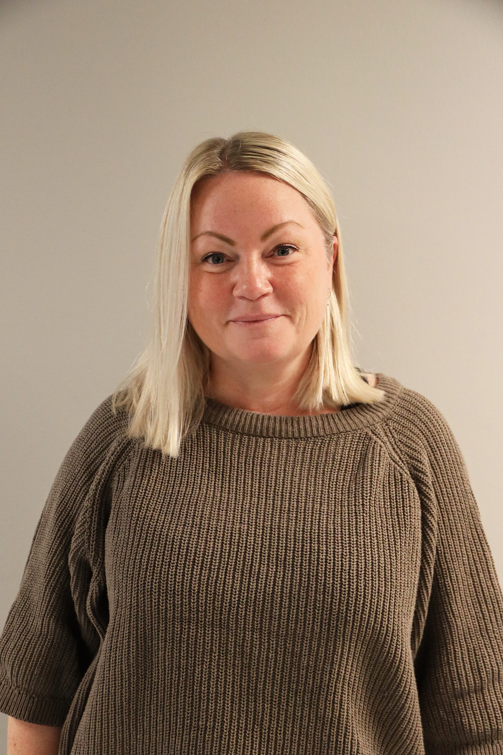 En kvinna med rakt blont hår och en brun stickad tröja står mot en vanlig ljus vägg och tittar in i kameran med ett neutralt uttryck.