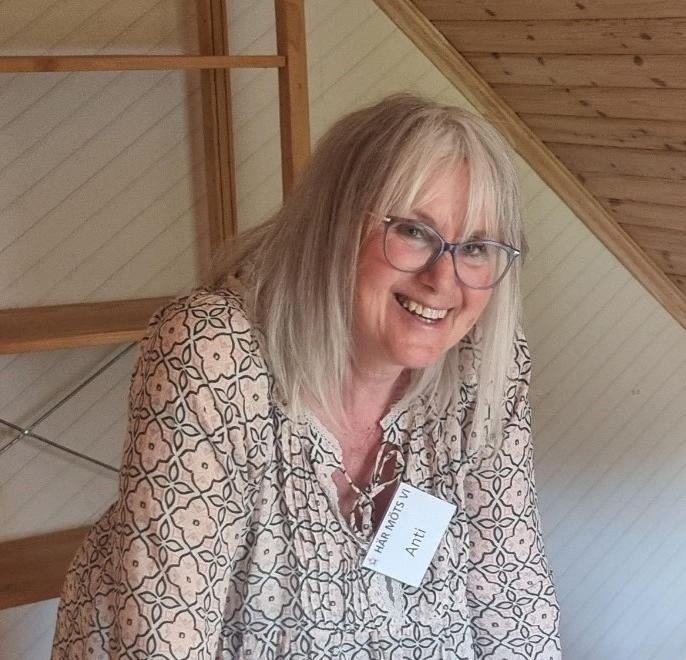 En person med grått hår och glasögon ler, iklädd mönstrad skjorta och namnskylt i ett rum med trädetaljer och en trappa.