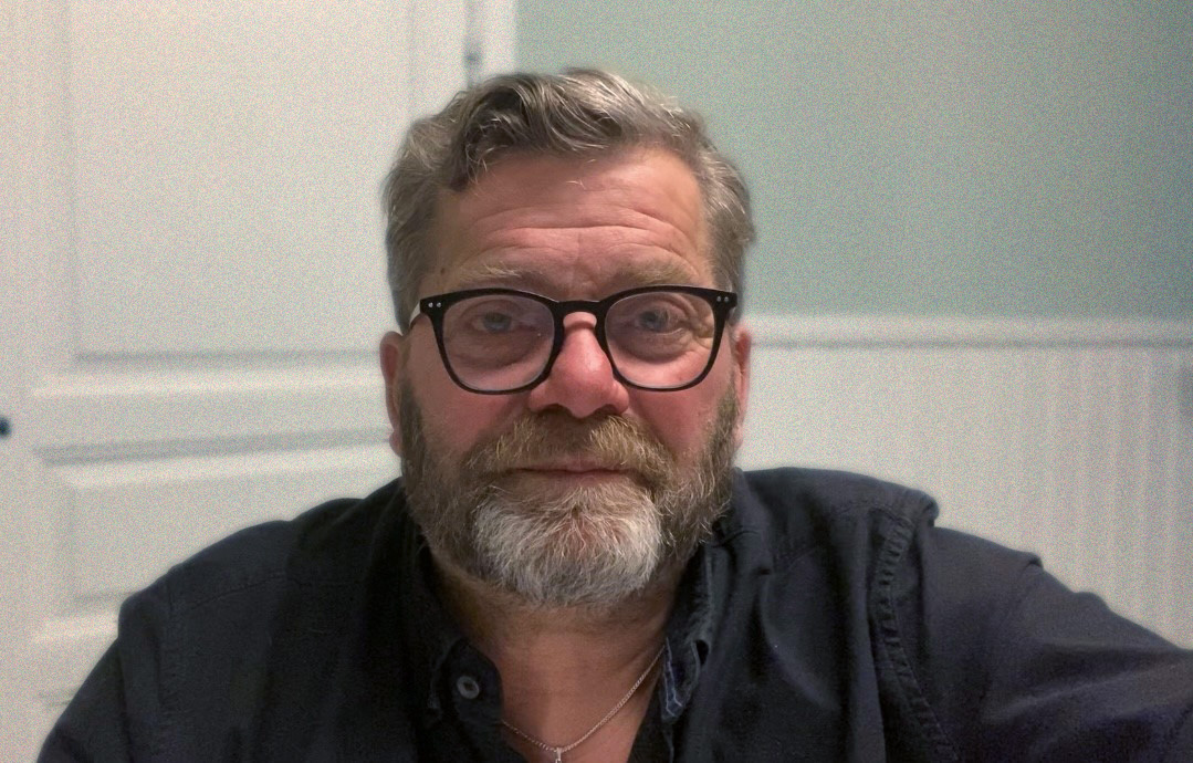 En person med grått hår, skägg och glasögon sitter inomhus och tittar på kameran med ett neutralt uttryck.