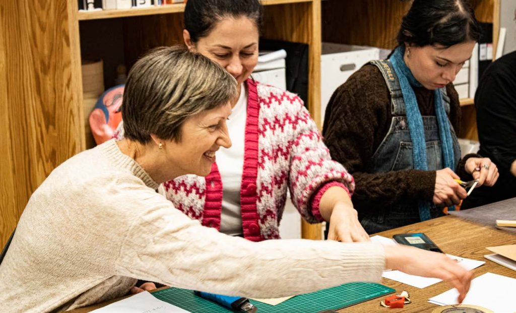 en grupp kvinnor som står runt ett träbord och utövar hantverk.