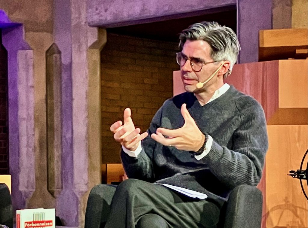 En man med glasögon, klädd i mörk tröja och vit skjorta, talar och gestikulerar med händerna under en sittande diskussion. Han har ett mikrofonheadset på sig.