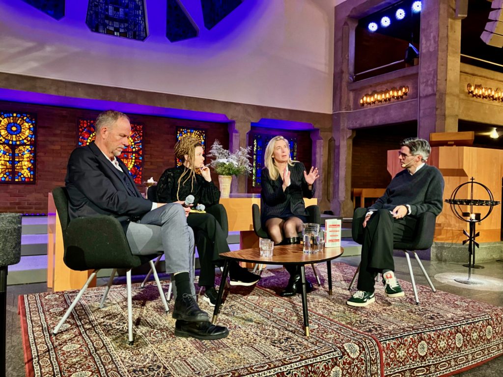 Fyra personer sitter på scenen i en paneldiskussion i en modern lokal med målade glasfönster och sitter runt ett litet bord med böcker och vattenglas.