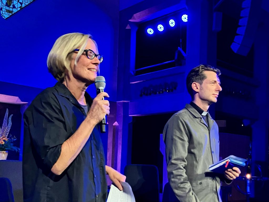 Två personer står på en scen i blått ljus; den ena håller i en mikrofon och papper, den andra håller i en anteckningsbok.