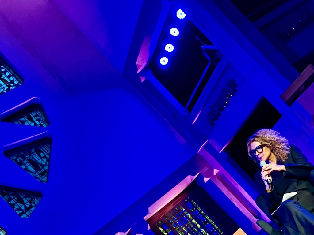 En person med lockigt hår talar i en mikrofon medan han sitter på en scen med blå belysning och geometriska glasmålningar i bakgrunden.