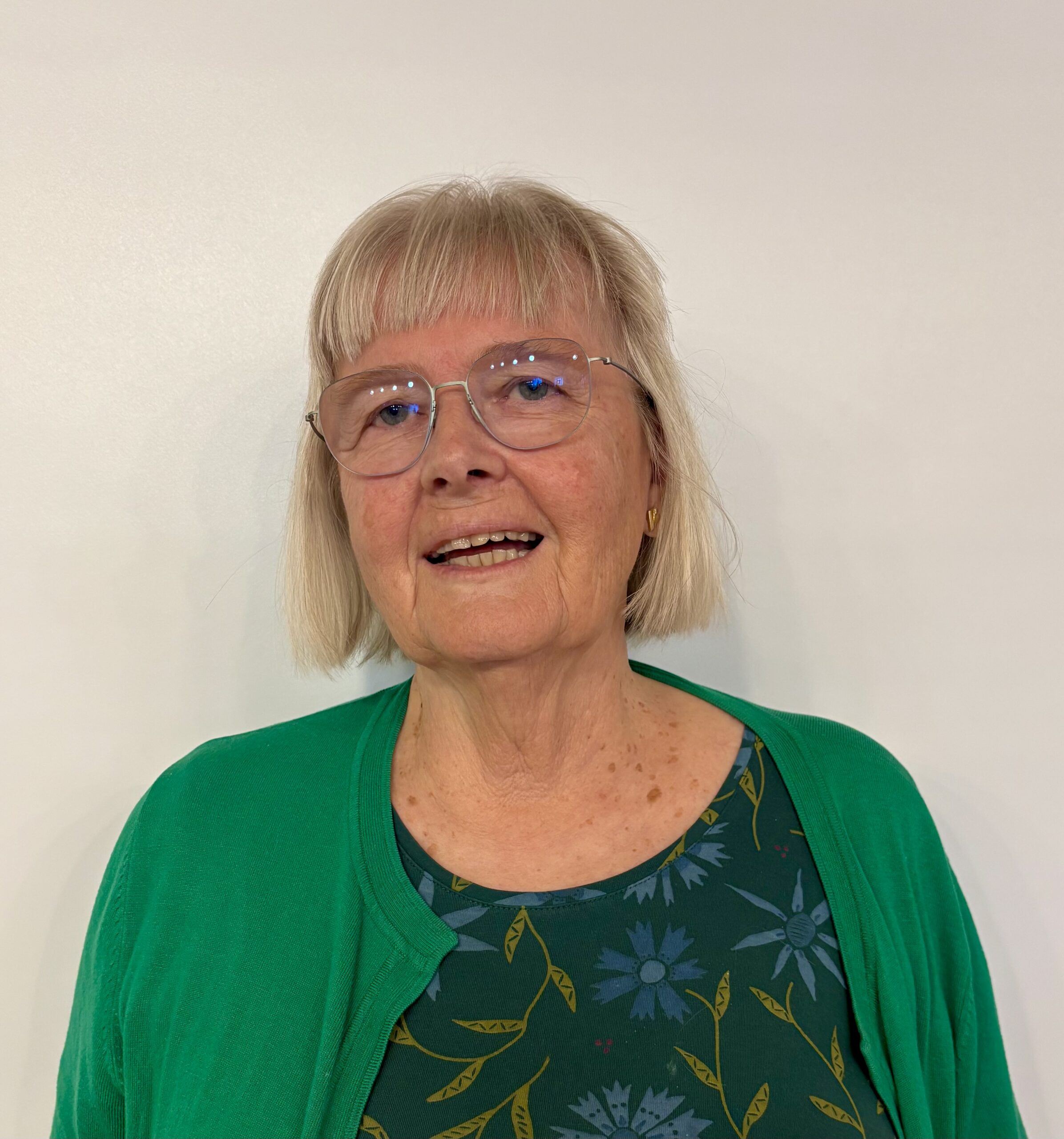 Äldre kvinna med kort grått hår, glasögon och grön tröja ler mot en ljus bakgrund.