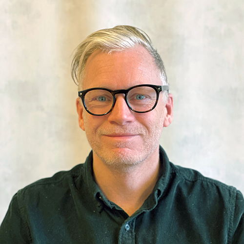 En person med kort blont hår, bär glasögon och mörk skjorta med knappar, ler mot kameran mot en ljus, strukturerad bakgrund.