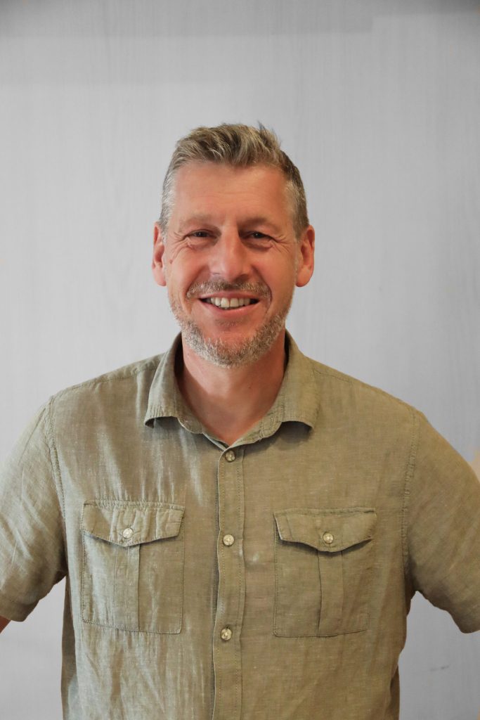 En leende man med kort grått hår och skägg, klädd i en ljusgrön skjorta med krage, står mot en vanlig ljus bakgrund.