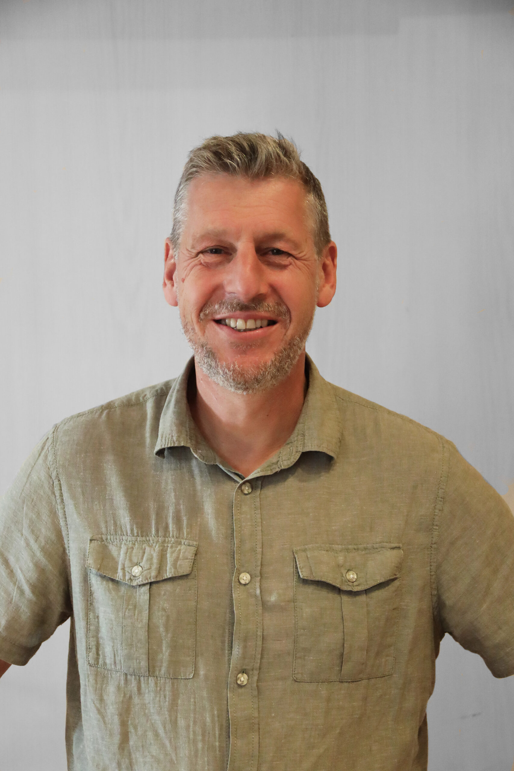 En leende man med kort grått hår och skägg, klädd i en ljusgrön skjorta med krage, står mot en vanlig ljus bakgrund.