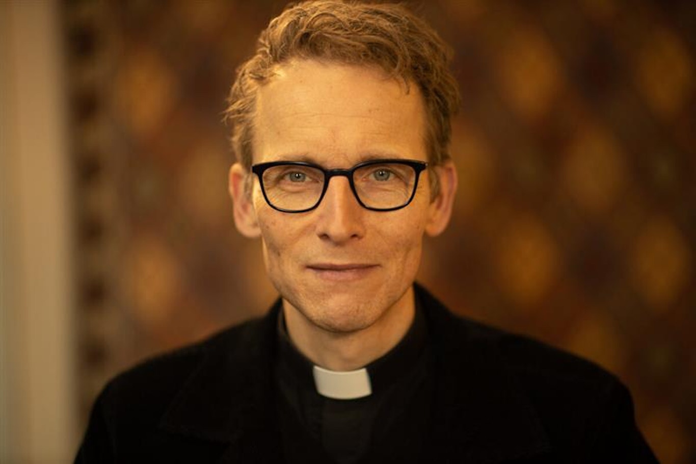 En man med glasögon och kontorskrage ler mjukt framför en suddig mönstrad bakgrund.