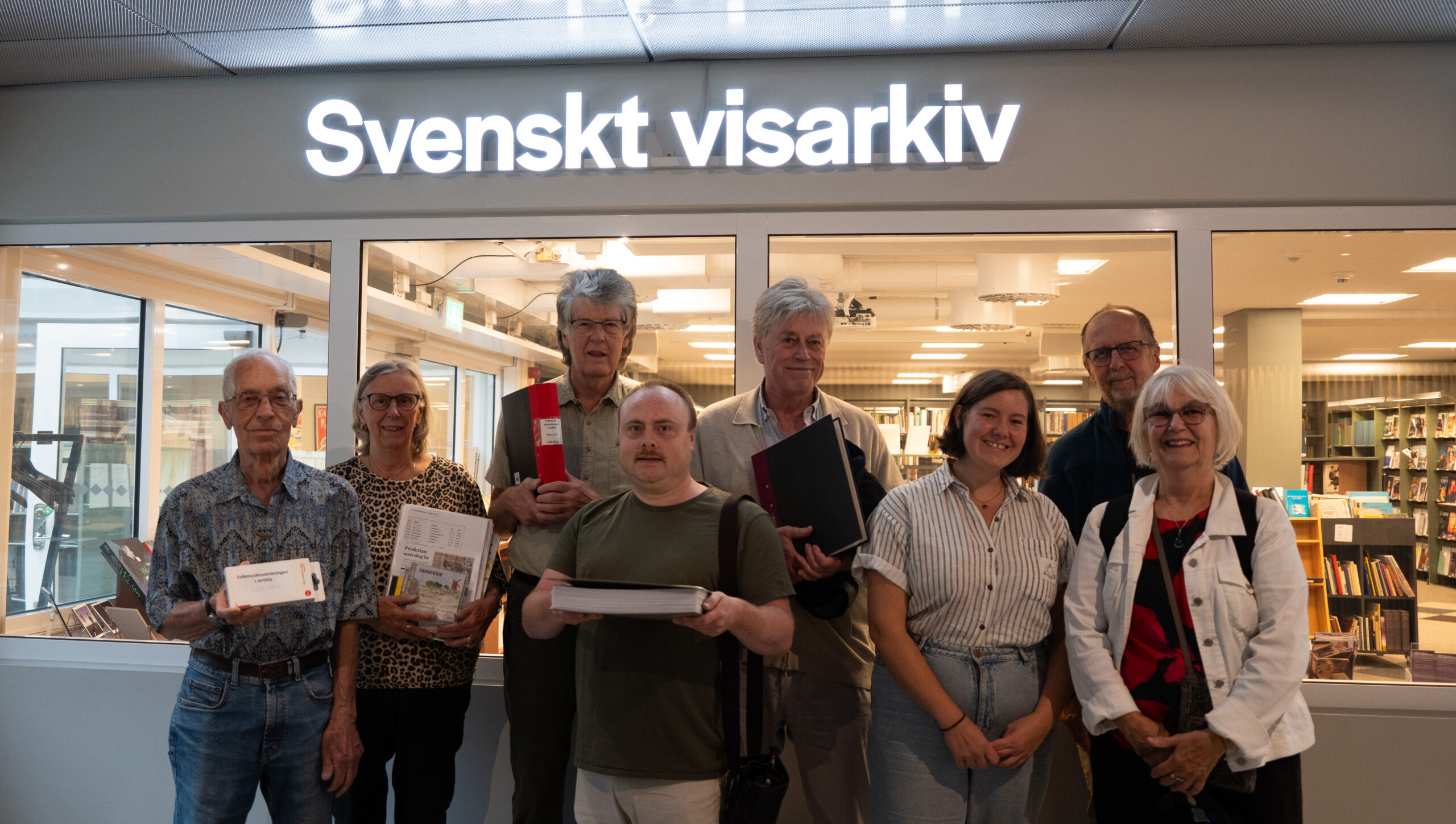 En grupp på åtta personer poserar framför ett fönster med skylten "Svenskt visarkiv", några med mappar och dokument i händerna.
