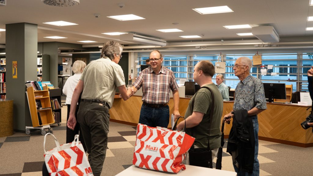 Fem män samtalar och skakar hand i en biblioteksmiljö, med rödvita MAKI-påsar på ett bord i förgrunden.