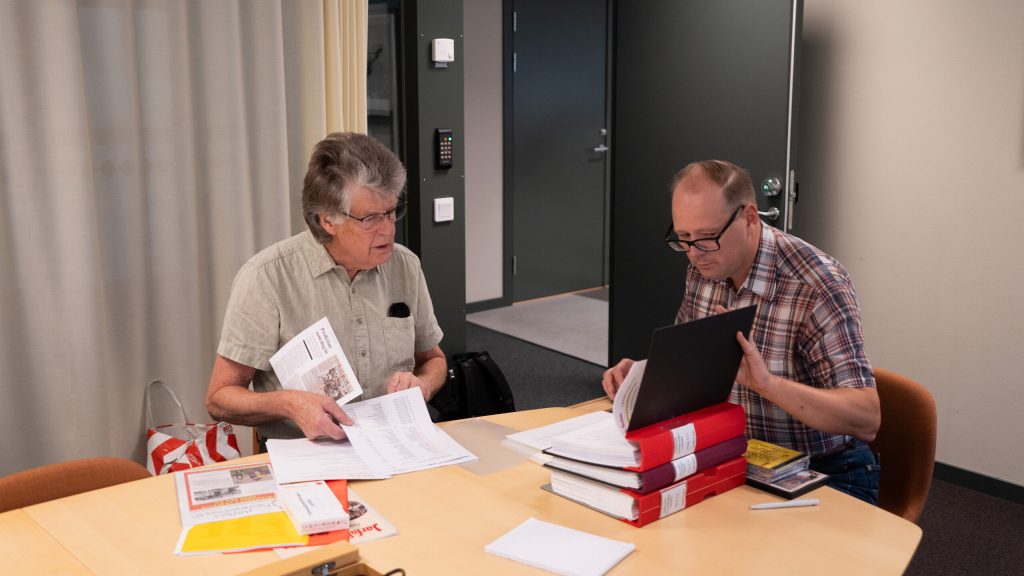 Två män sitter vid ett bord med papper, pärmar och en bärbar dator och ser ut att diskutera eller granska dokument i en kontorsmiljö.