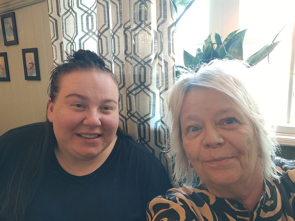 Två kvinnor sitter inomhus vid ett fönster och ler mot kameran. Den ena har mörkt hår och bär en svart skjorta, den andra har kort, ljust hår och en mönstrad topp.