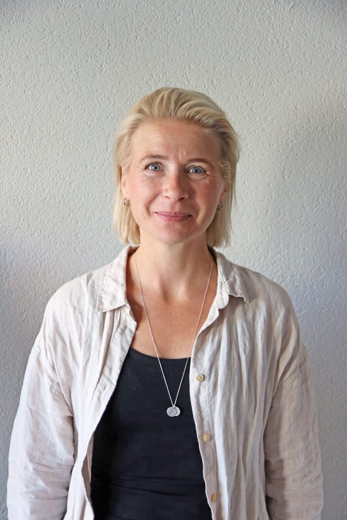 En kvinna med kort blont hår, klädd i en ljus skjorta med knappar över en svart topp, står framför en ljusgrå vägg med textur och tittar in i kameran.