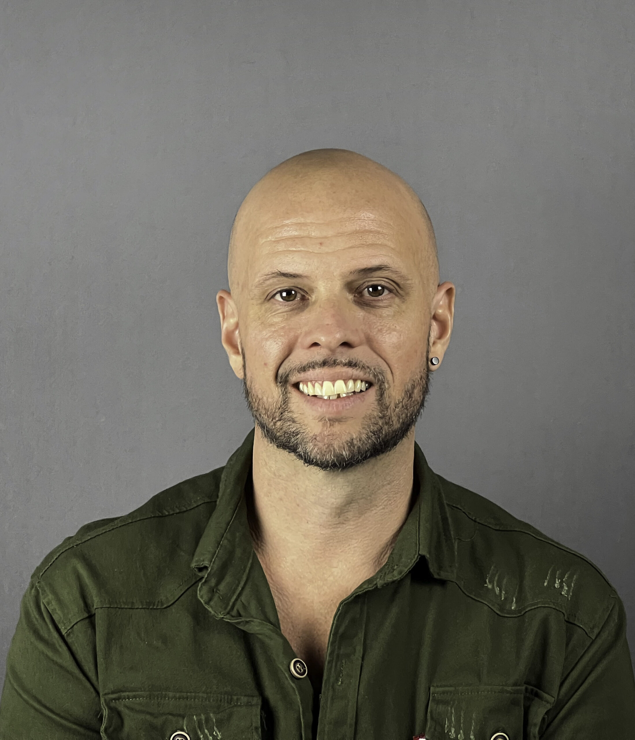 Skallig man med trimmat skägg och grön skjorta med knappar ler mot en grå bakgrund.