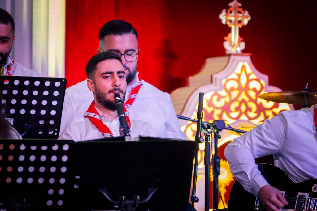 Musiker i vita skjortor uppträder på scenen med röd och guld bakgrund; en spelar klarinett, en annan gitarr.