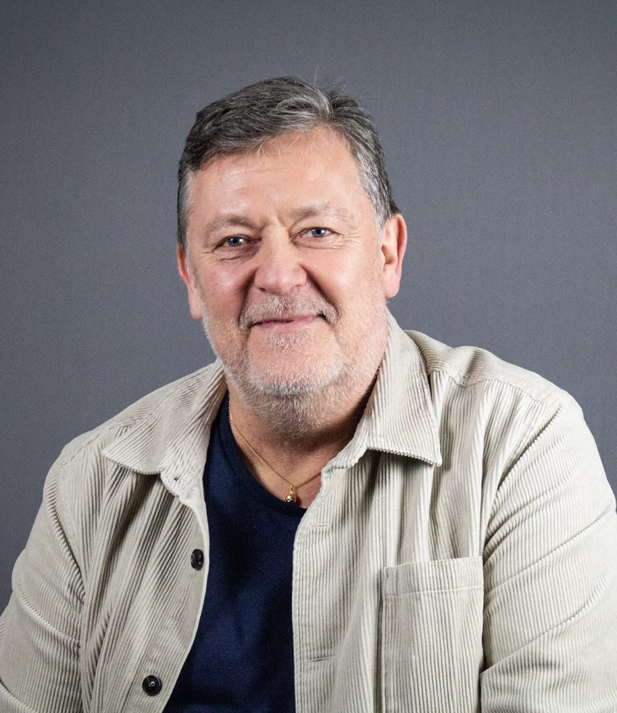 En man med kort grått hår, klädd i randig beige jacka och mörk skjorta, ler framför en vanlig grå bakgrund.