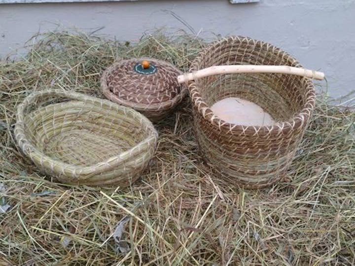 Tre handvävda halmkorgar, en med handtag, en med lock och en öppen, placerade på en hög med hö.