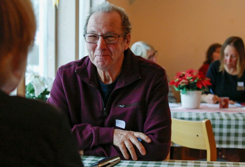 Leende äldre man i glasögon sitter vid ett bord på ett mysigt café med andra människor i bakgrunden.