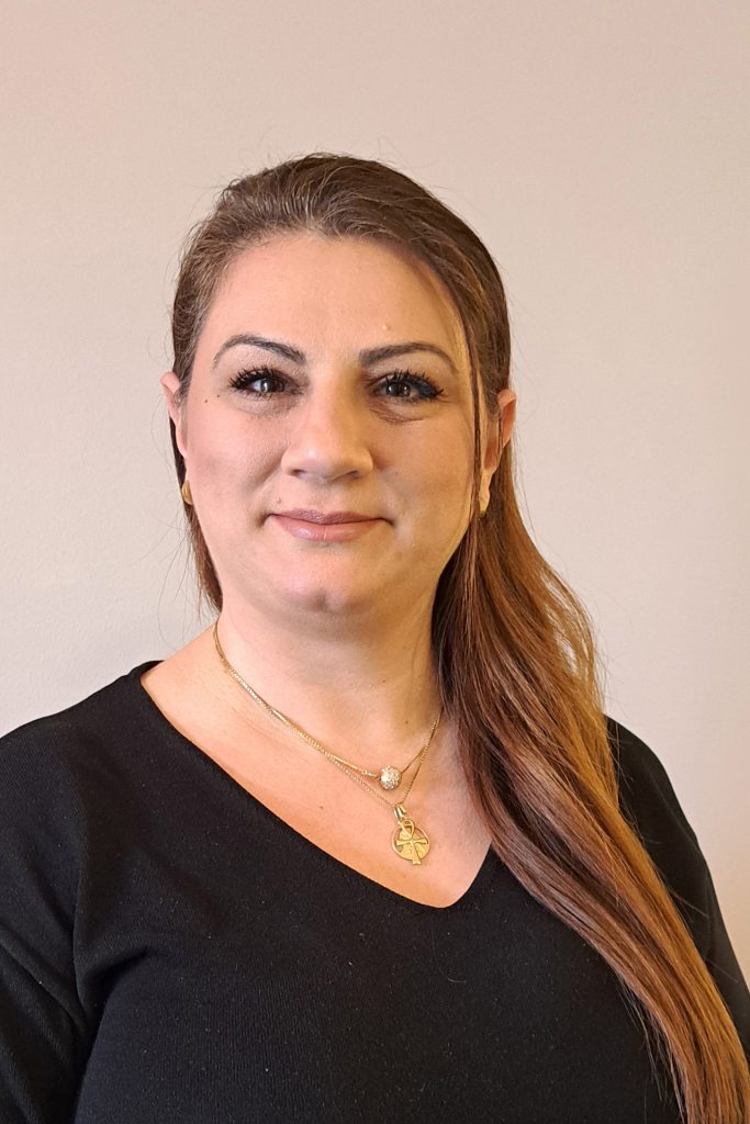 Kvinna med långt brunt hår i svart topp, bär guldsmycken och ler mot kameran mot en ljus bakgrund.