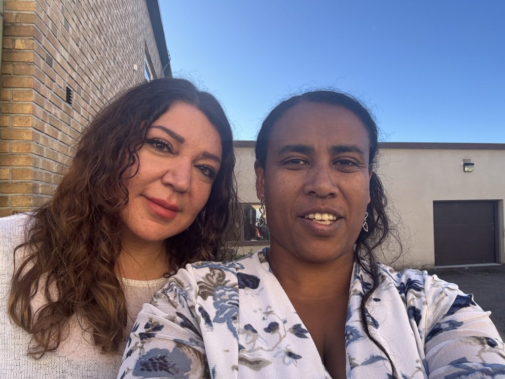 Två kvinnor poserar för en selfie utomhus nära tegel- och beigefärgade byggnader under en klarblå himmel.