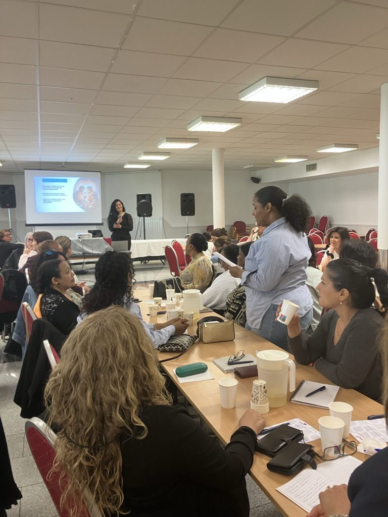En kvinna presenterar för en grupp som sitter vid bord med kaffekoppar, anteckningsböcker och en projicerad bild bakom henne.
