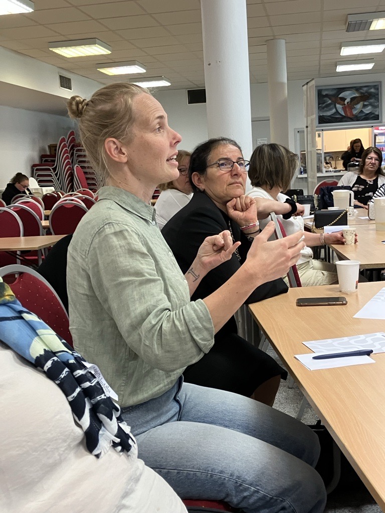 En kvinna talar på ett möte och gestikulerar med händerna, medan andra lyssnar runt ett bord med papper och koppar.