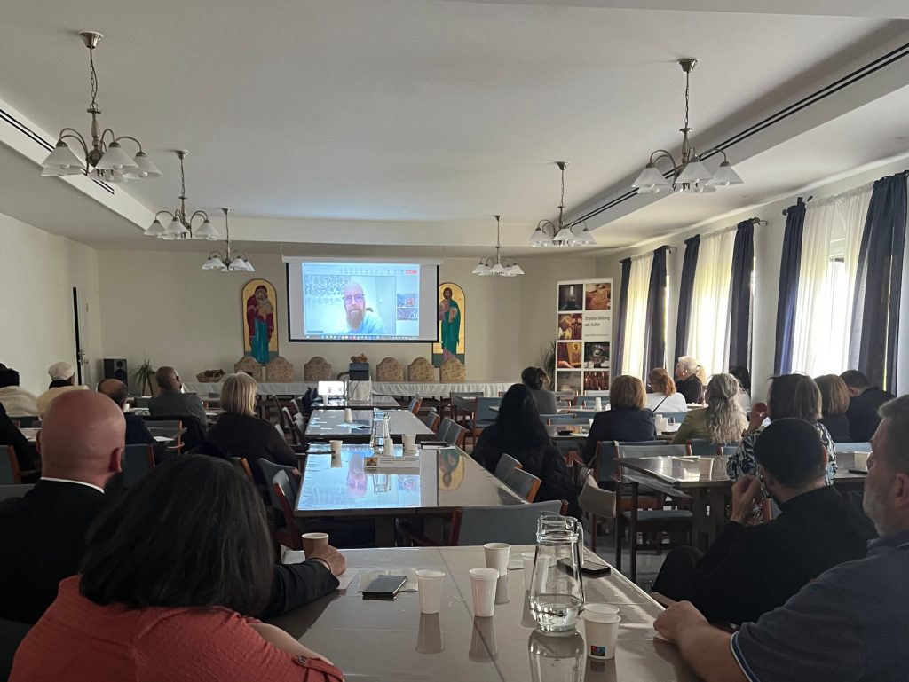 Personer som sitter i ett konferensrum och tittar på en talare i ett videosamtal som projiceras på en skärm.