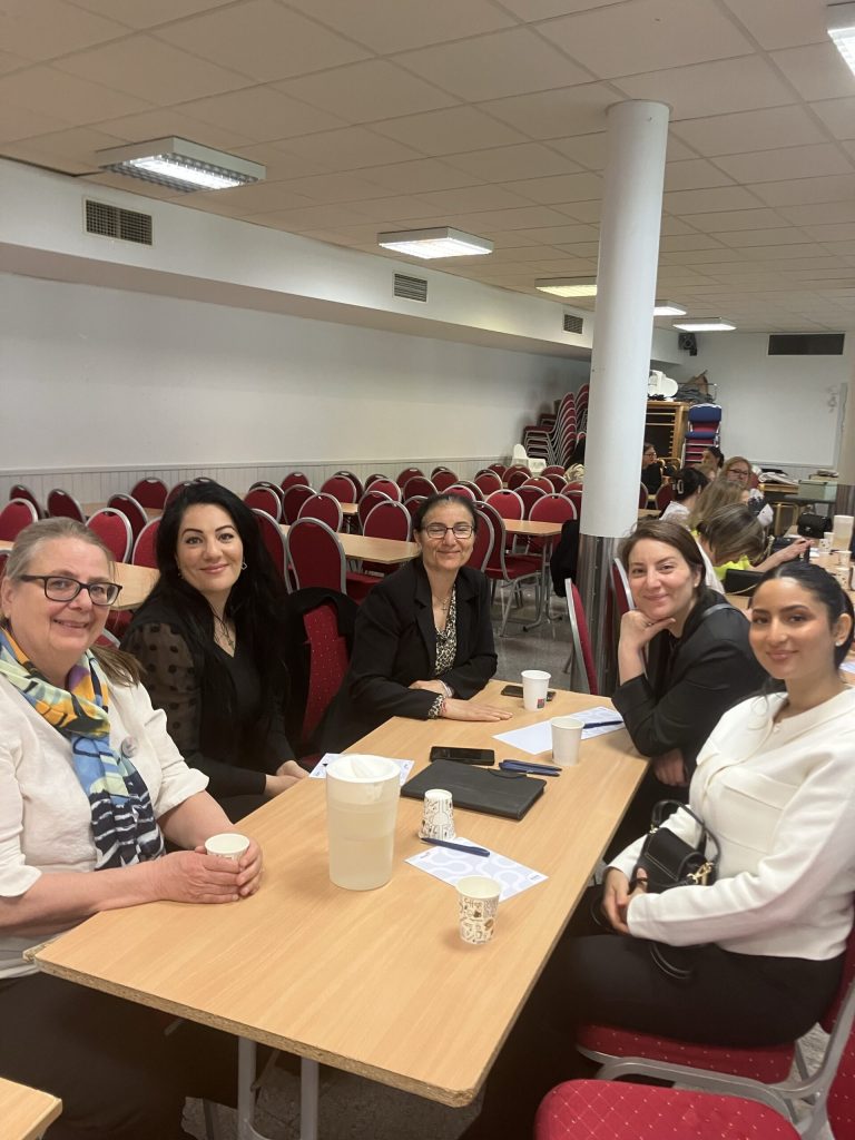 Fem kvinnor sitter runt ett bord i ett konferensrum, ler mot kameran och har kaffekoppar på bordet.