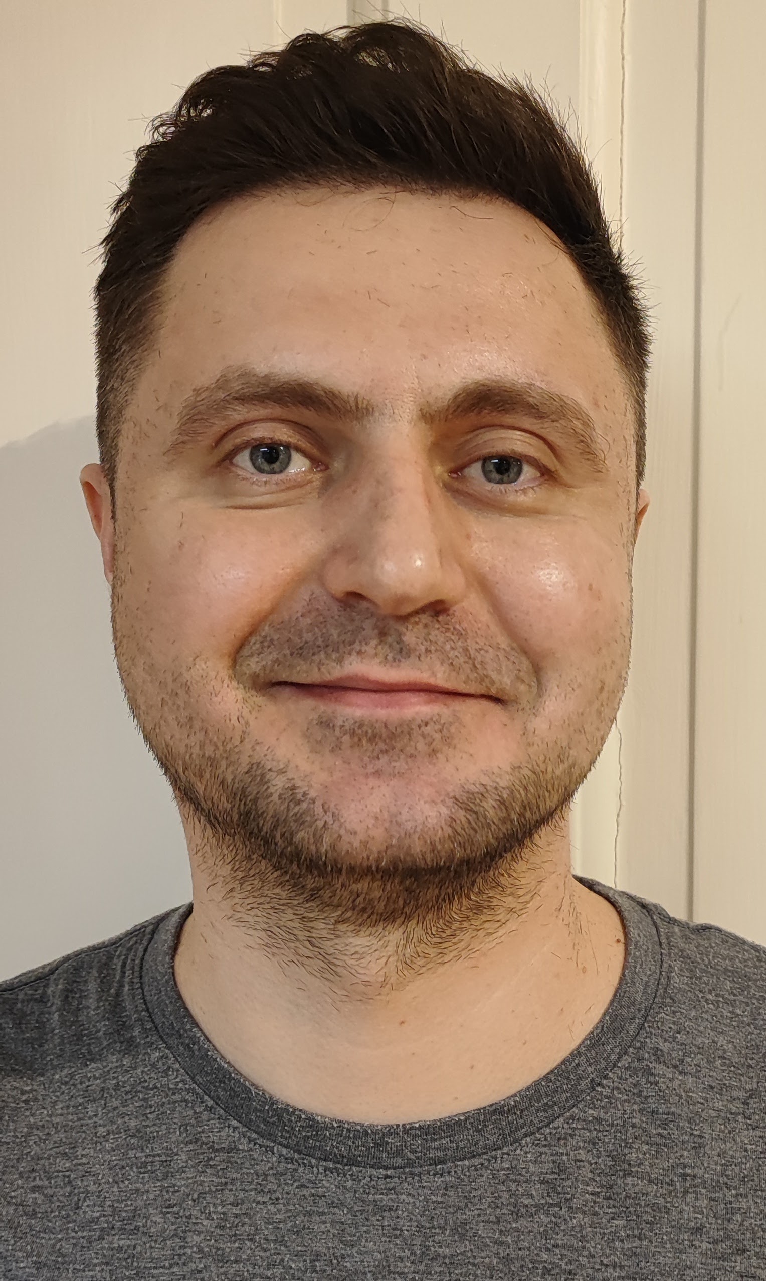 En man med kort mörkt hår och ljust skägg, klädd i en grå t-shirt, ler lätt mot en neutral bakgrund.