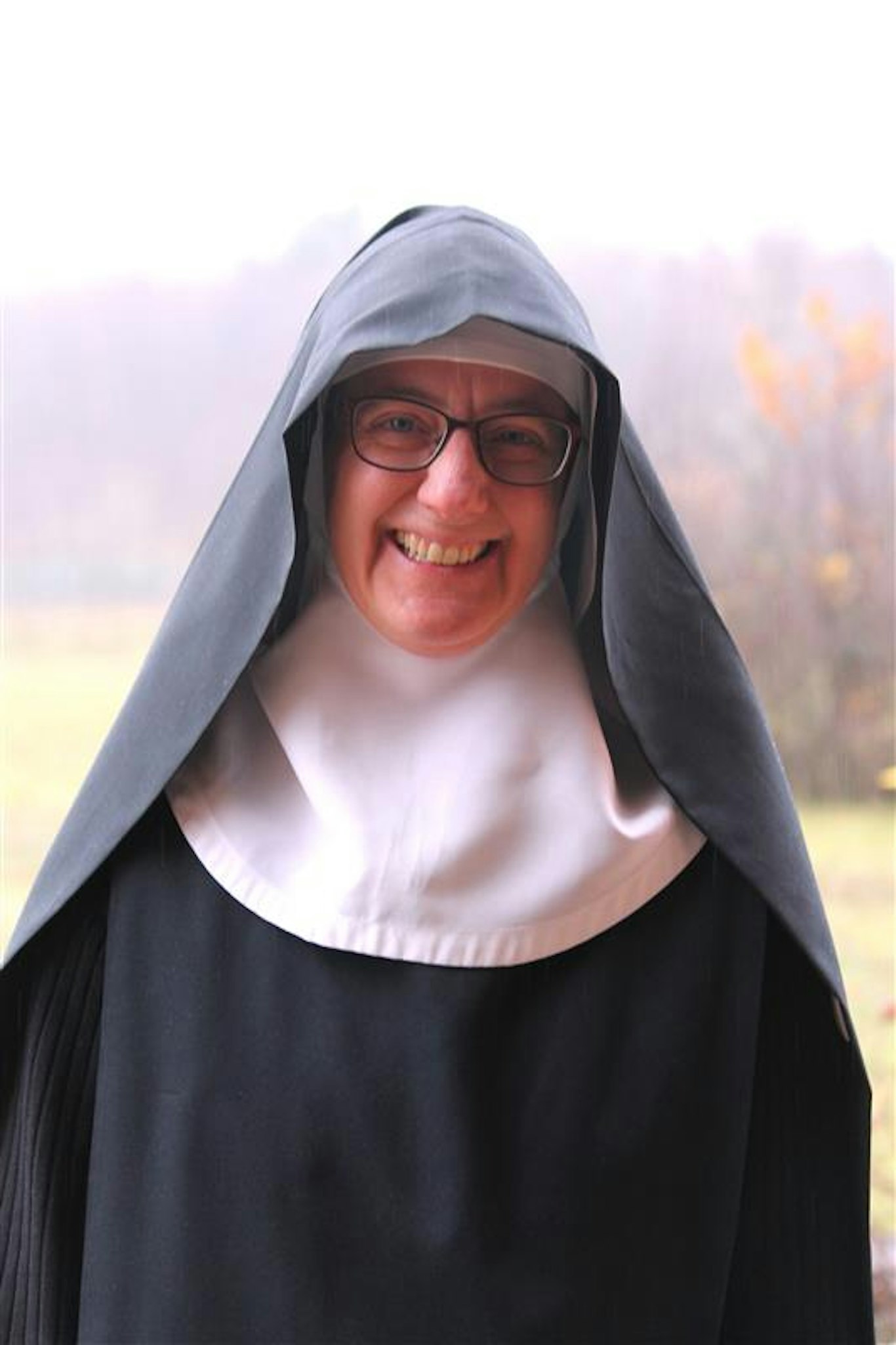 En leende nunna i svartvit klädedräkt och glasögon står utomhus en molnig dag.