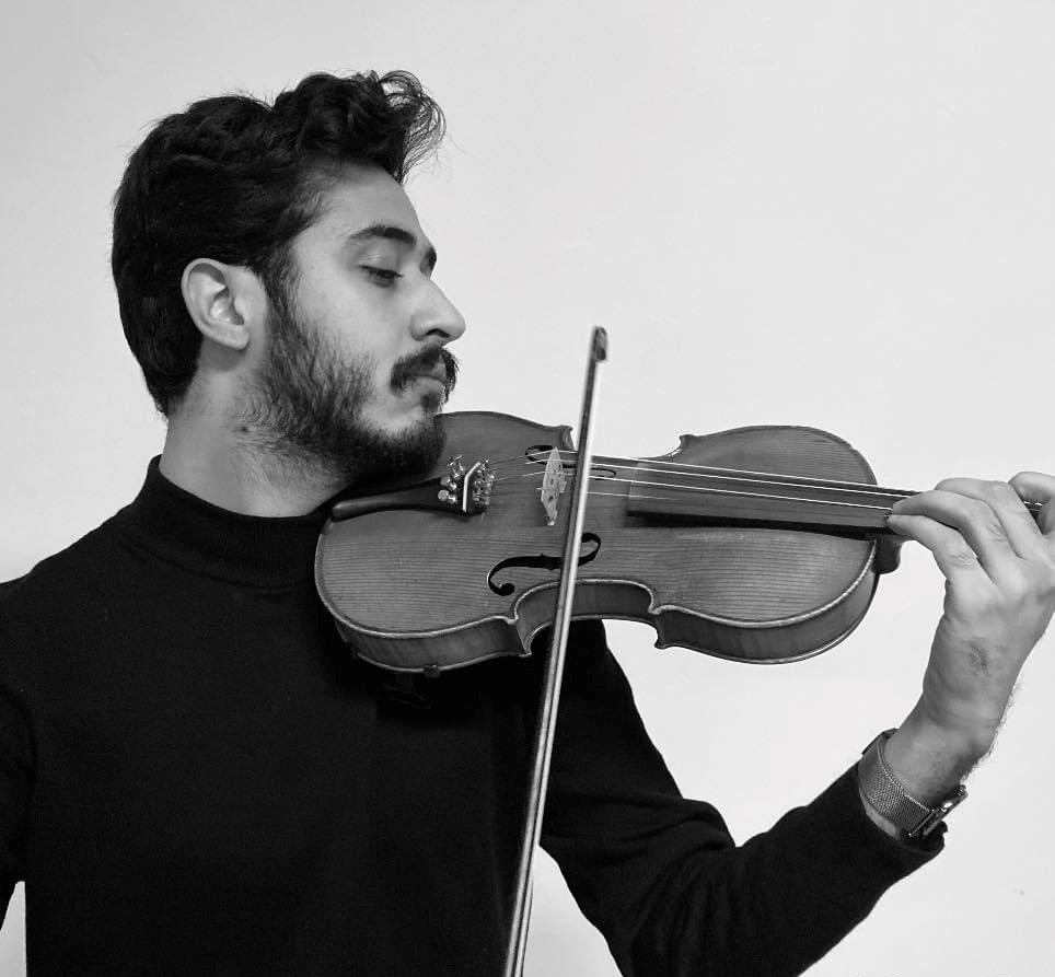 En person spelar fiol med ett fokuserat uttryck, klädd i mörk turtleneck. Bakgrunden är enkel.